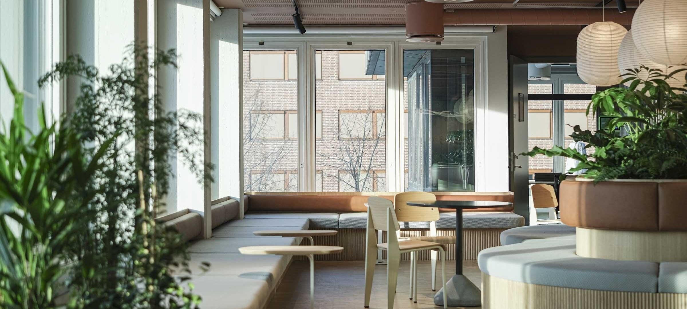 Resepsjonsområde med spesialtegnet sittegruppe med grønne planter i midten og belysning som henger fra taket. Andre små sittegrupper plassert rundt. Vinduer langs fasade med spesiallaget benk under rekken med vinduer. Foto