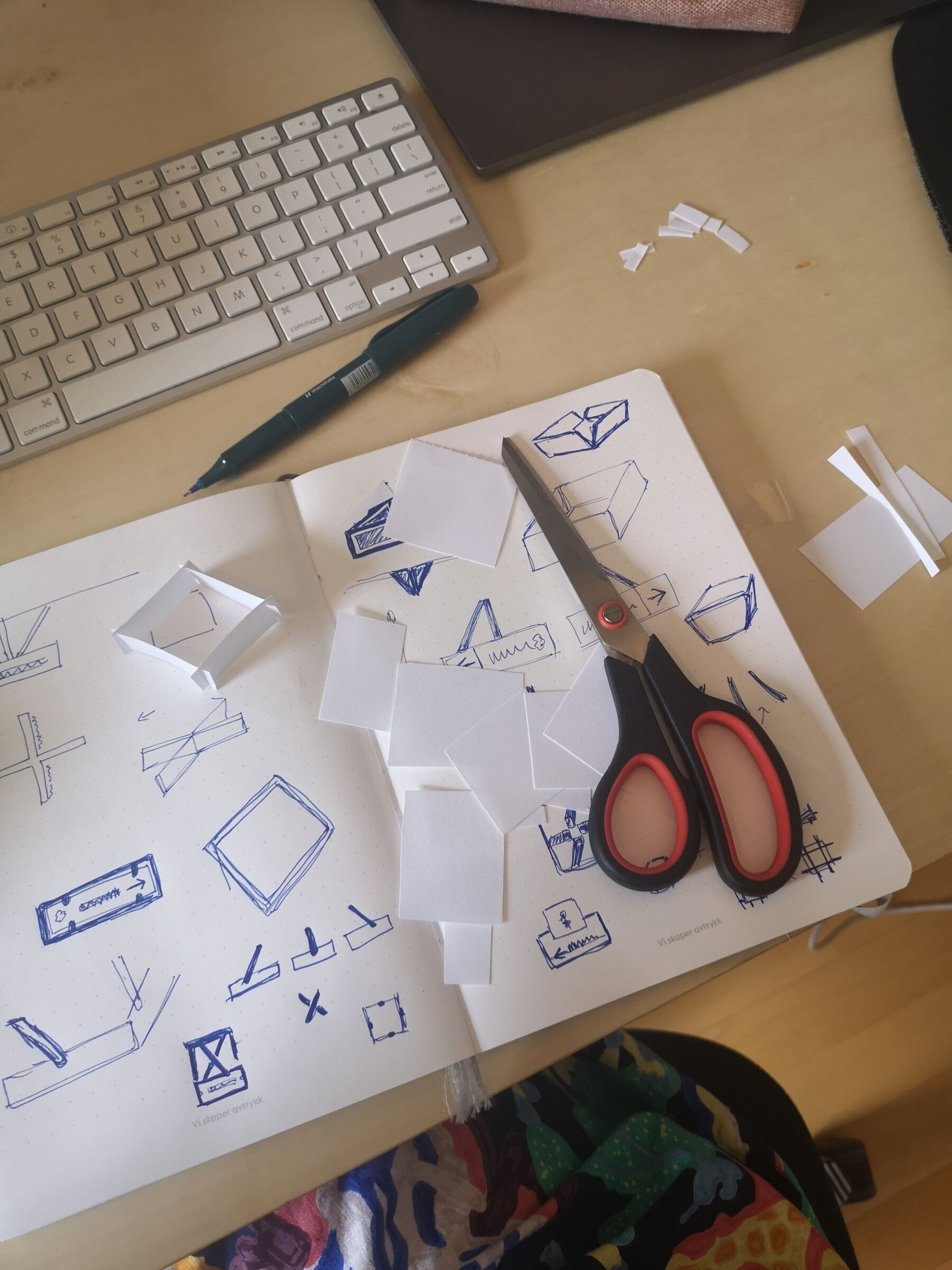 Skissebok med tegning av grafisk arbeid. Saks, tastatur og penn plassert sammen med skissebok. Foto